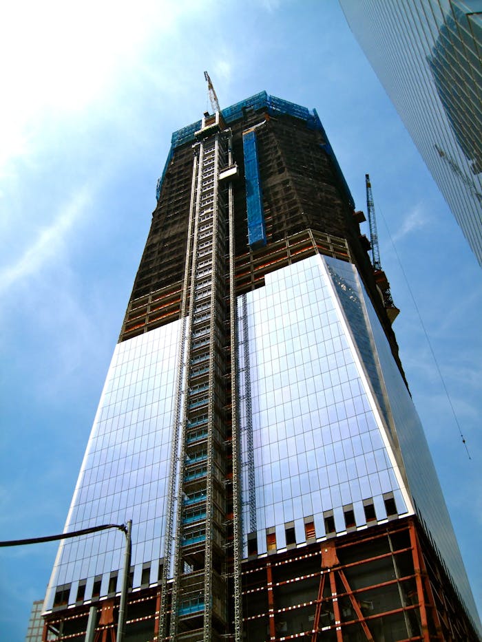 A towering modern skyscraper under construction with cranes and reflective glass against a clear blue sky.